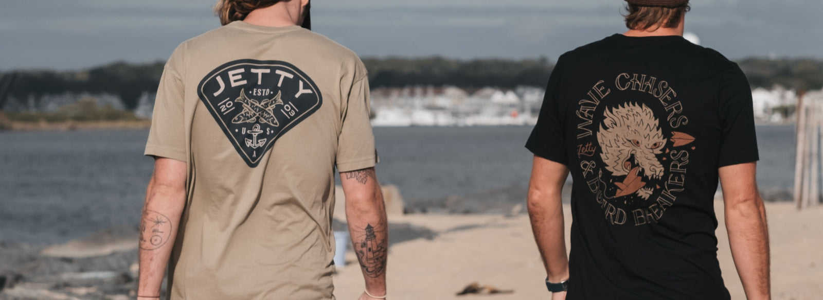 Two men wearing graphic tees walking on the jetty with their back towards us, showing us the back design of the shirts